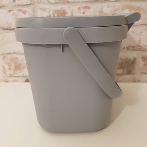 May include: A gray plastic bucket with a handle and a lid. The bucket is sitting on a white surface.