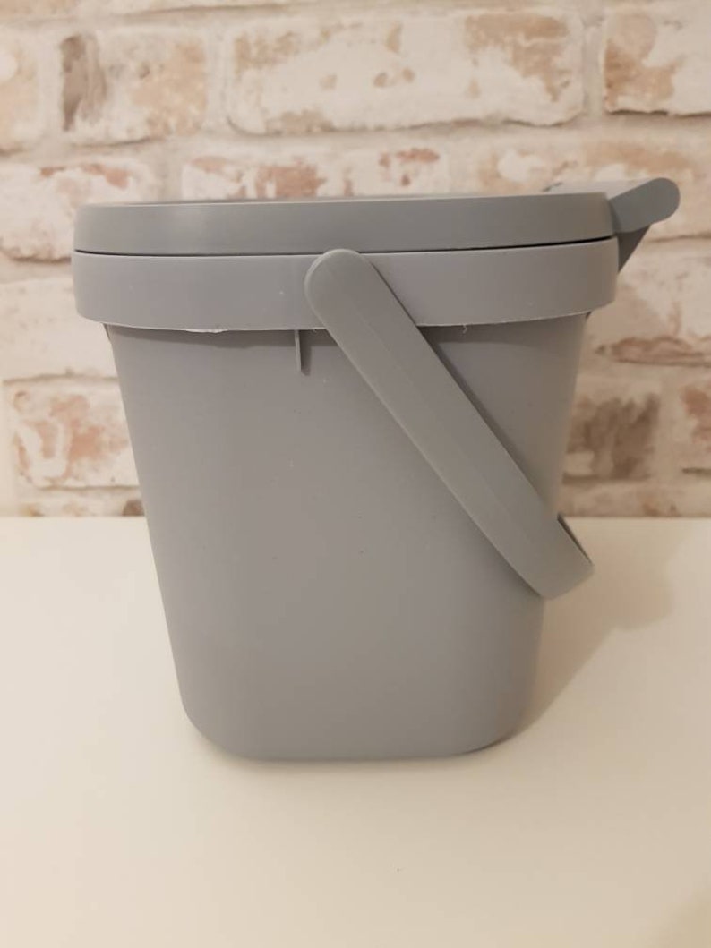 May include: A gray plastic bucket with a handle and a lid. The bucket is sitting on a white surface.