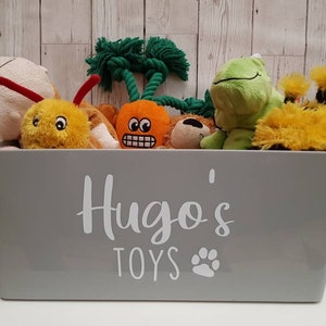 Op de afbeelding: Een grijze opbergbak met de tekst "Hugo's Toys" en een pootprint. De bak is gevuld met verschillende pluche speelgoed, waaronder een groene kikker, een gele bij, een wortel en een bruine beer.