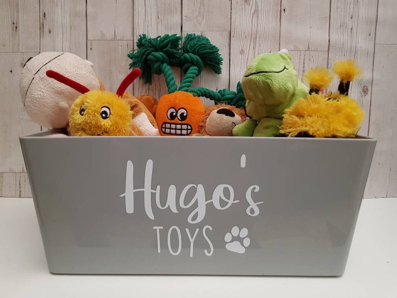 Op de afbeelding: Een grijze opbergbak met de tekst "Hugo's Toys" en een pootprint. De bak is gevuld met verschillende pluche speelgoed, waaronder een groene kikker, een gele bij, een wortel en een bruine beer.