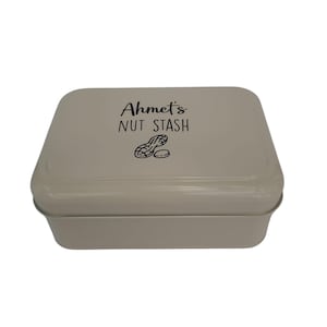 May include: A light beige metal storage tin with a lid. The lid has the text "Ahmet's NUT STASH" in black script, and a peanut graphic. The tin is rectangular and designed for storing nuts or snacks, with a matching light beige colour.