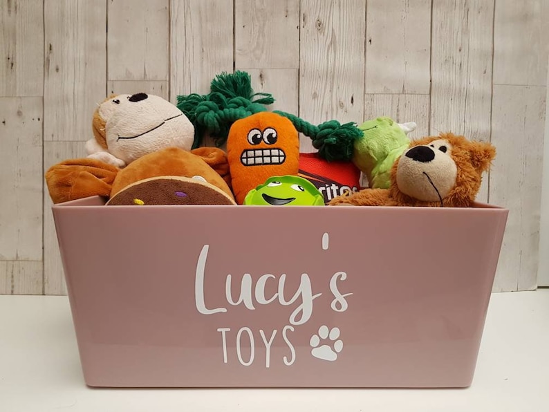 Op de afbeelding: Een roze opbergbak met de tekst "Lucy's Toys" en een pootprint. De bak is gevuld met knuffels, waaronder een bruine aap, een groen-wit wezen, een speelgoed in de vorm van een wortel en een bruine beer.