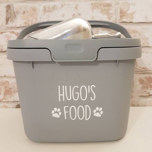May include: Gray plastic pet food container with a scoop inside. The container has a white label that says "Hugo's Food" with paw prints on either side.