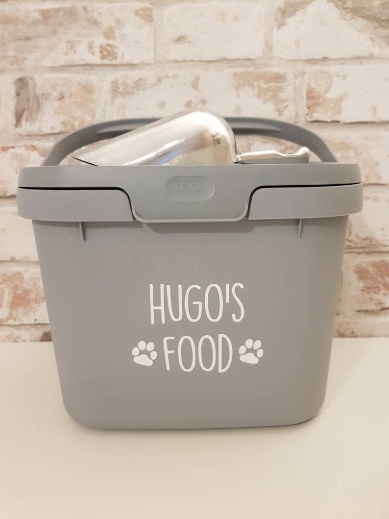 May include: Gray plastic pet food container with a scoop inside. The container has a white label that says "Hugo's Food" with paw prints on either side.