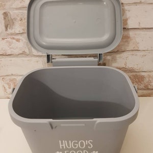 May include: Gray plastic pet food container with a lid. The container has a white paw print design and the text "HUGO'S FOOD" printed on the front.