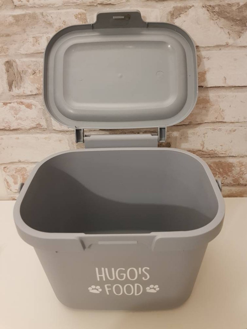 May include: Gray plastic pet food container with a lid. The container has a white paw print design and the text "HUGO'S FOOD" printed on the front.