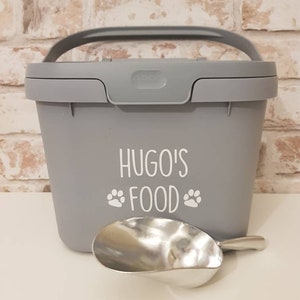 May include: Gray plastic pet food container with a handle and a scoop. The container has the text "Hugo's Food" with paw prints on the front.
