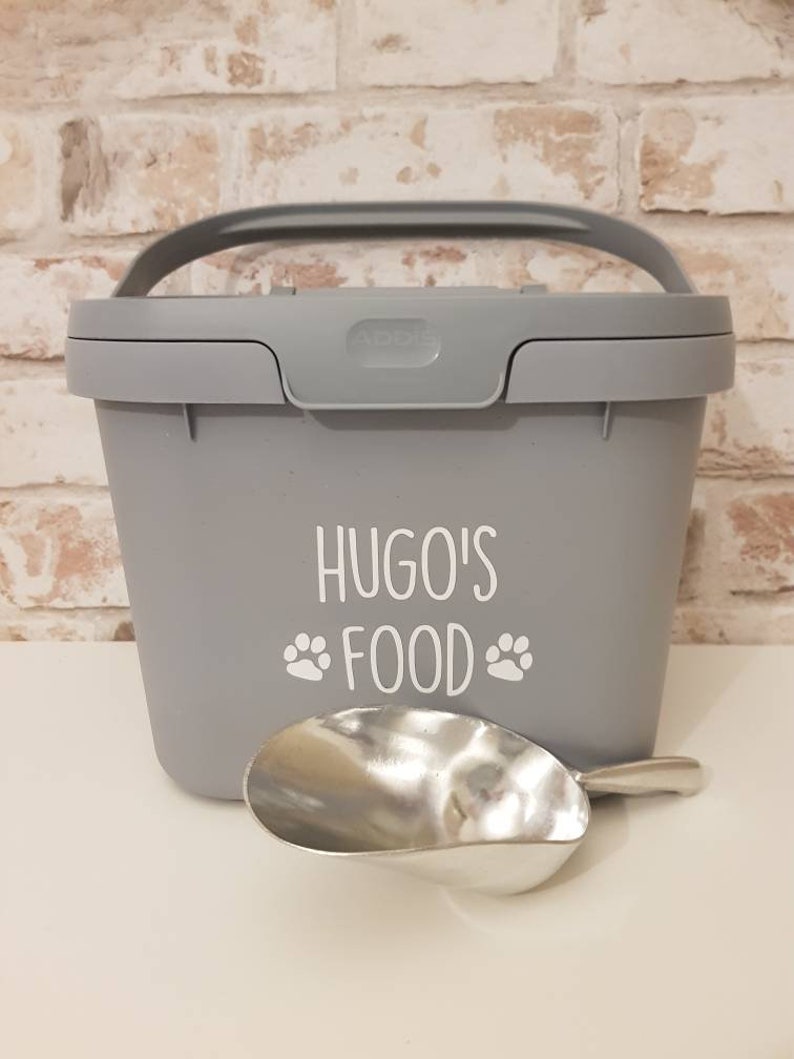 May include: Gray plastic pet food container with a handle and a scoop. The container has the text "Hugo's Food" with paw prints on the front.