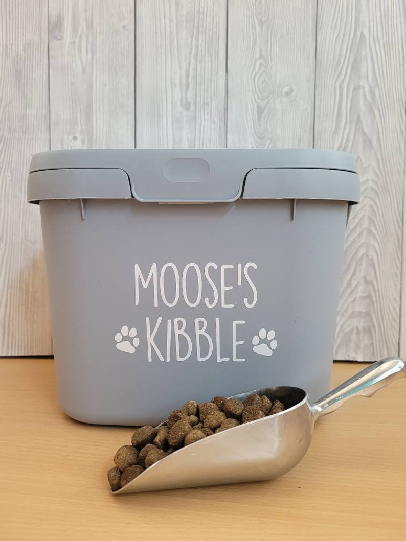 May include: Gray plastic pet food container with a white label that says "Moose's Kibble" with paw prints. A silver scoop is filled with brown kibble.