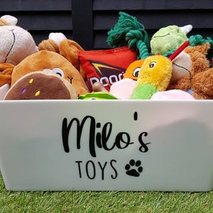 Op de afbeelding: Een witte plastic opbergbak met de woorden "Milo's Toys" en een pootprint op de voorkant. De bak is gevuld met een verscheidenheid aan knuffels, waaronder een aap, een bruine hond, een groene dinosaurus, een gele wortel en een groen-gele slang.