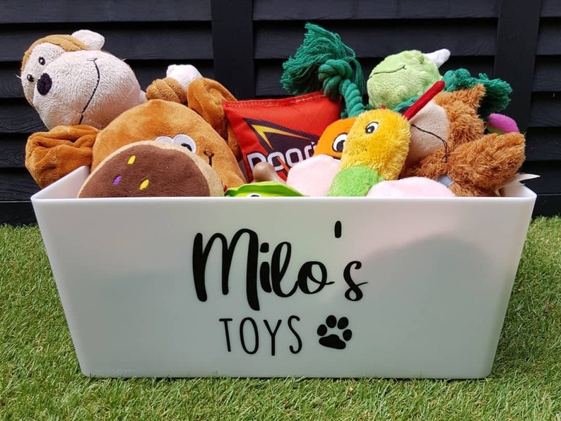 Op de afbeelding: Een witte plastic opbergbak met de woorden "Milo's Toys" en een pootprint op de voorkant. De bak is gevuld met een verscheidenheid aan knuffels, waaronder een aap, een bruine hond, een groene dinosaurus, een gele wortel en een groen-gele slang.
