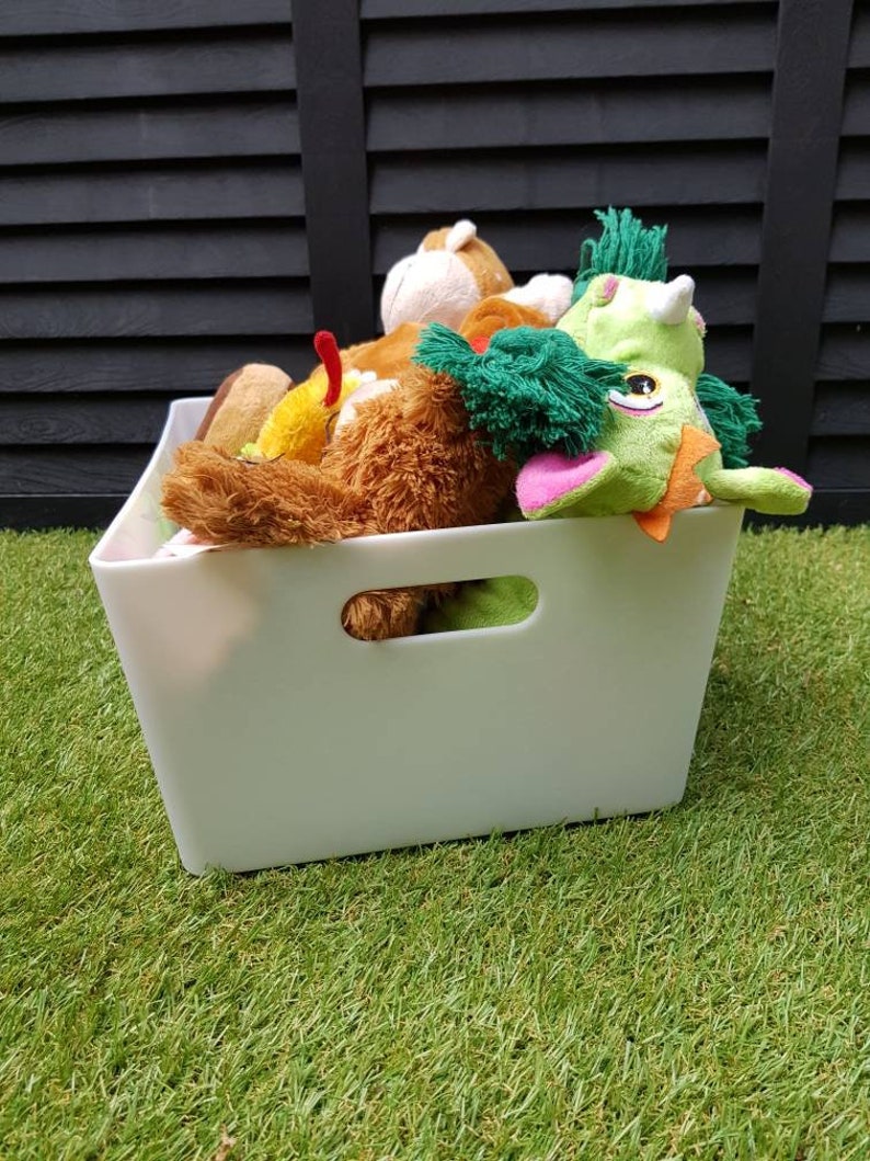 Op de afbeelding: Een witte plastic opbergbox met een handvat, gevuld met verschillende pluche speelgoed, waaronder een groene en roze draak, een bruine beer en een bruine en witte aap.