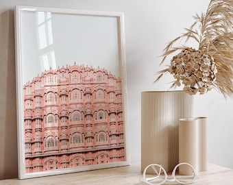 May include: A framed print of a pink building with many windows and arches. The print is hanging on a wall above a wooden desk with two drawers. A vase with dried flowers and a pair of white glasses are on the desk.