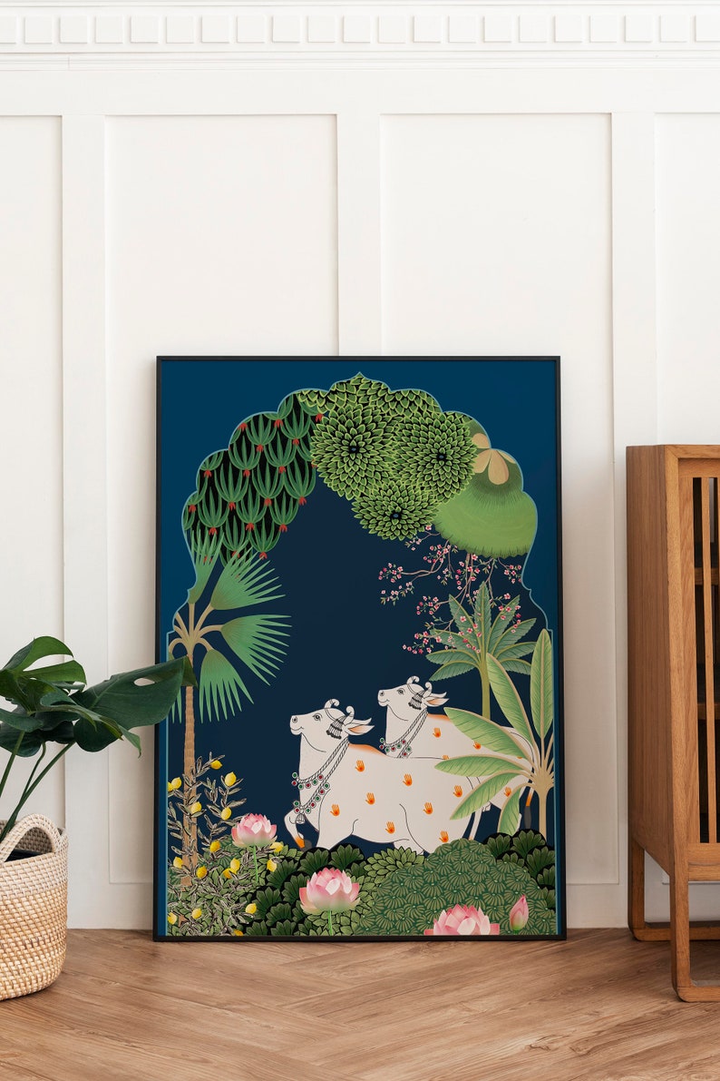 May include: A framed illustration of two white cows with orange spots walking through a lush green forest. The cows are adorned with floral garlands and bells. The background is a deep blue with a traditional archway design.