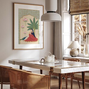 May include: A framed illustration of a woman sitting under a palm tree with pink lotus flowers and a peacock. The illustration is in a gold frame and is hanging on a white wall. The wall is behind a white dining table with a white marble top and two wicker chairs.