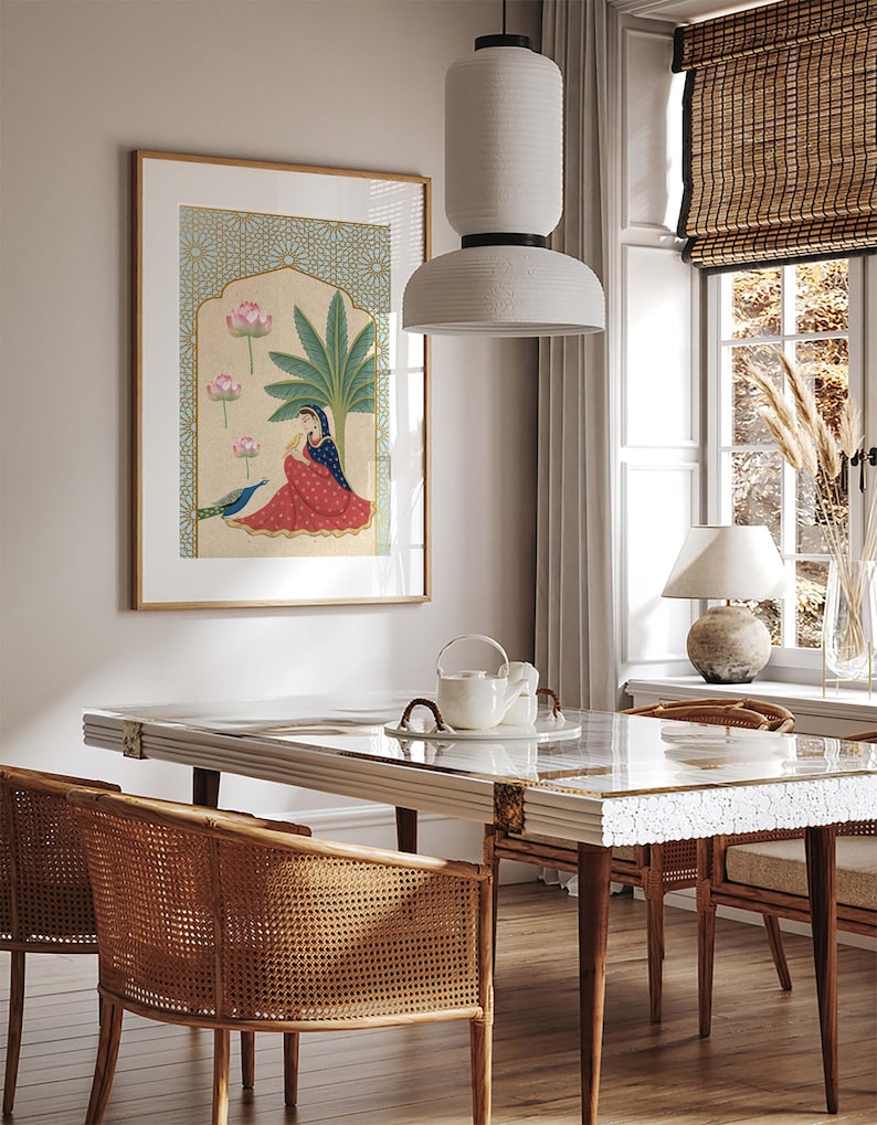 May include: A framed illustration of a woman sitting under a palm tree with pink lotus flowers and a peacock. The illustration is in a gold frame and is hanging on a white wall. The wall is behind a white dining table with a white marble top and two wicker chairs.