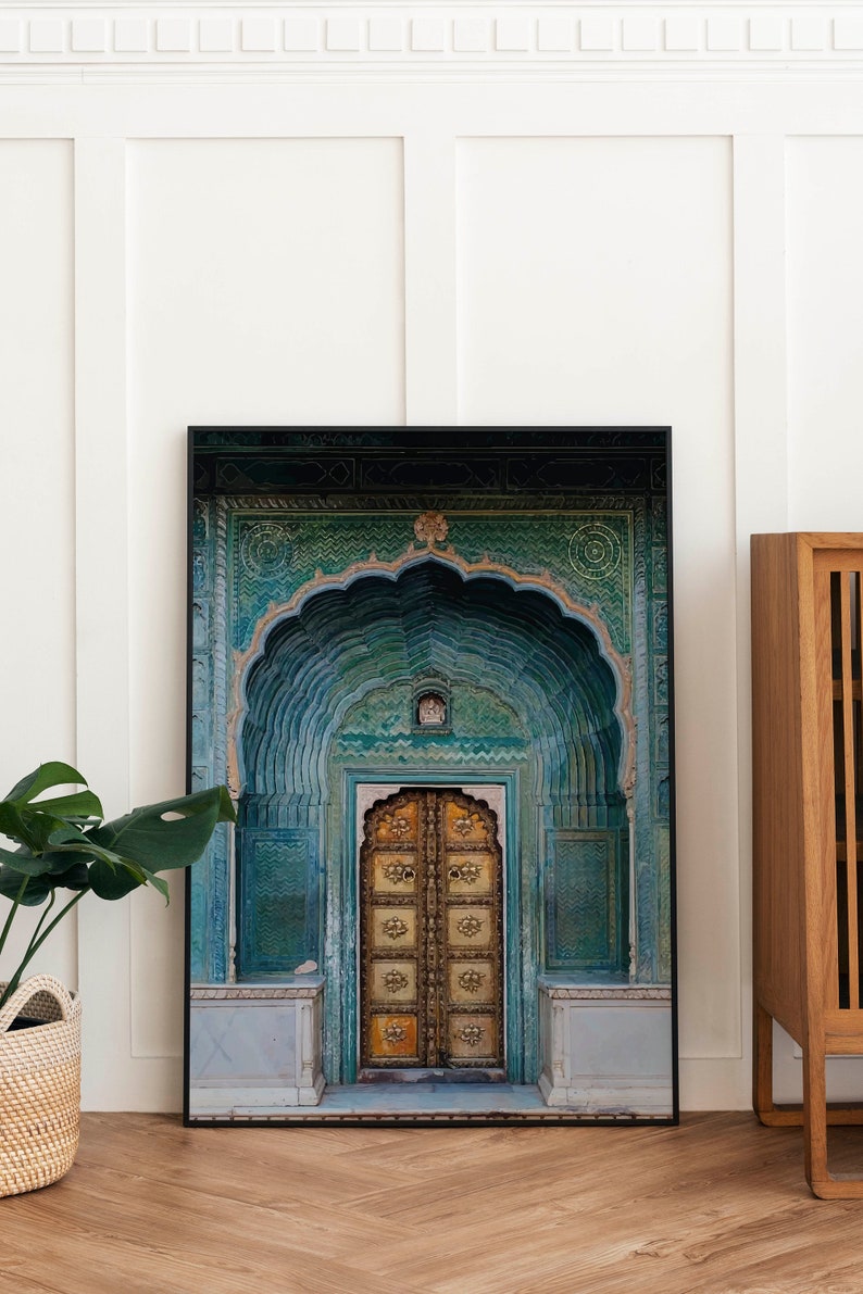 May include: Framed art print featuring a detailed image of an ornate, arched doorway. The door is a warm gold color, set within a teal and turquoise facade. The artwork is displayed on a wooden floor, with a plant and a wooden cabinet in the background.