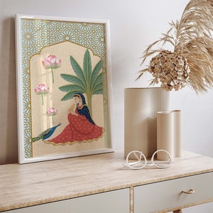 May include: A framed illustration of a woman in a red and blue dress sitting with a peacock. The illustration is set against a background of a palm tree and pink lotus flowers. The frame has a gold and blue geometric pattern.