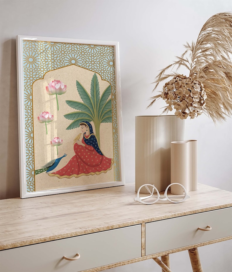 May include: A framed illustration of a woman in a red and blue dress sitting with a peacock. The illustration is set against a background of a palm tree and pink lotus flowers. The frame has a gold and blue geometric pattern.