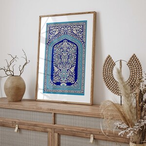 Puede incluir: Una impresi&oacute;n enmarcada de una alfombra azul y blanca con intrincados dise&ntilde;os florales. La alfombra est&aacute; enmarcada en un marco de madera natural y est&aacute; colgada en una pared blanca sobre un armario de madera.