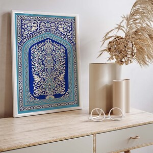 Puede incluir: Una impresi&oacute;n enmarcada de una alfombra azul y blanca con intrincados dise&ntilde;os florales. La alfombra se exhibe en una mesa de madera con dos jarrones de cer&aacute;mica blanca y un par de gafas redondas blancas.