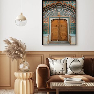 May include: A framed print of an ornate, golden arched doorway with teal and orange decorative elements. The print is displayed above a brown sofa with decorative pillows. A side table with a glass vase and pampas grass is next to the sofa.