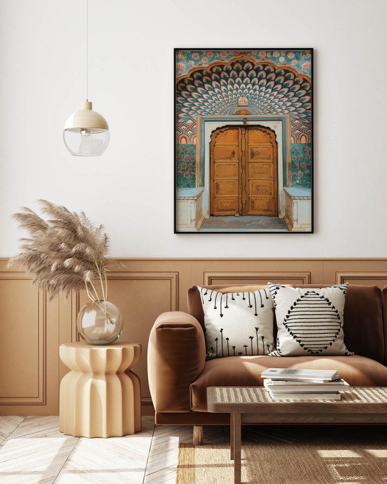 May include: A framed print of an ornate, golden arched doorway with teal and orange decorative elements. The print is displayed above a brown sofa with decorative pillows. A side table with a glass vase and pampas grass is next to the sofa.