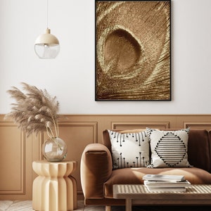 May include: A brown velvet sofa with two throw pillows, one with a black and white geometric pattern and the other with a white and black arrow pattern. The sofa is in front of a wall with a framed abstract print in shades of brown. A small, round, beige side table with a vase of dried pampas grass sits next to the sofa.