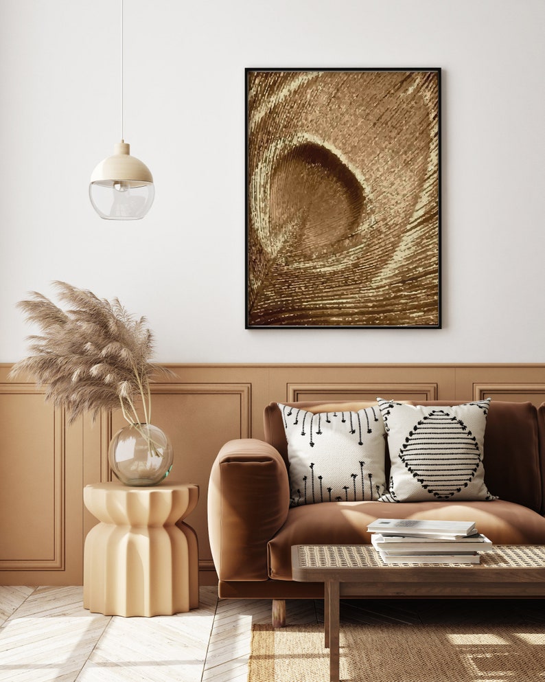 May include: A brown velvet sofa with two throw pillows, one with a black and white geometric pattern and the other with a white and black arrow pattern. The sofa is in front of a wall with a framed abstract print in shades of brown. A small, round, beige side table with a vase of dried pampas grass sits next to the sofa.