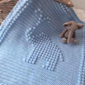 May include: A light blue, crocheted baby blanket with an elephant design. The blanket features a textured pattern and a border of bobbles. A small, gray crocheted elephant toy sits on the blanket.
