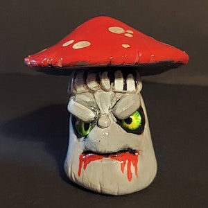 May include: A hand-painted ceramic mushroom figurine with a red cap and white spots. The mushroom has a grey face with green eyes and a dripping red mouth.