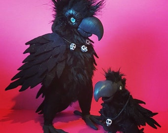 May include: Two black crow figurines with a punk rock aesthetic. The larger crow has a mohawk and a necklace with skull charms. The smaller crow has a necklace with a single skull charm.