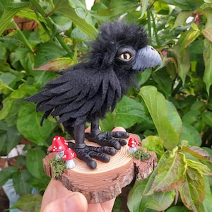 Può includere: Figurina artigianale di corvo nero con corpo soffice, becco grigio e occhi gialli. Si erge su una base di legno con accenti di funghi rossi e bianchi in miniatura. I piedi del corvo sono dettagliati.