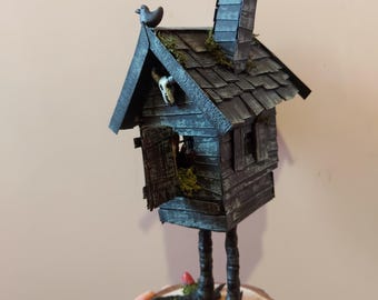 May include: A whimsical, dark-colored miniature house with a chimney, perched on chicken legs. The house features a small bird on the roof, a skull in the window, and sits on a wooden base with miniature skulls and mushrooms. The house is approximately 6 inches tall.