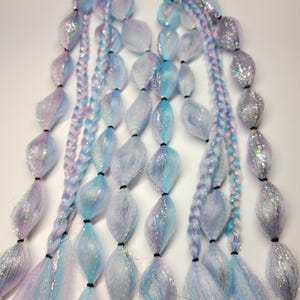 Puede incluir: Dos juegos de extensiones de cabello con mechones azules, rosas y blancos. Cada juego tiene una banda elástica negra en la parte superior y presenta una serie de cuentas azules y blancas iridiscentes.