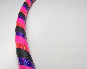Può includere: Un cerchio di hula a strisce rosa, nero e viola. Il cerchio è fatto di plastica e ha una finitura lucida.