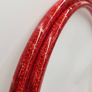 Puede incluir: Primer plano de dos aros rojos con purpurina. Los aros son curvos y tienen una superficie brillante y texturizada con purpurina incrustada. Los aros son de color rojo vibrante y parecen estar hechos de un material flexible.