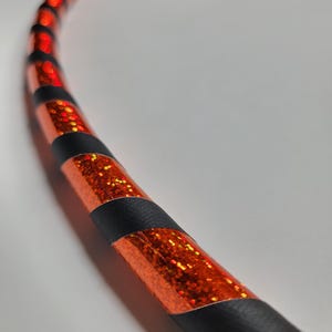 Pode incluir: Uma secção de um arco com um vibrante acabamento de glitter laranja, acentuado por faixas pretas. O arco é curvo, sugerindo uma forma circular, e é provavelmente usado para fitness ou brincadeiras. O design é apelativo e moderno.