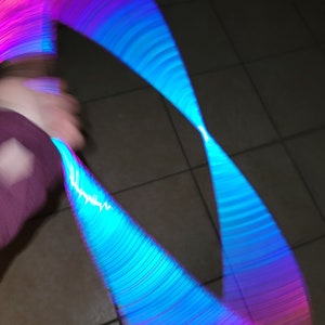 Op de afbeelding: Een blauwe en roze lichtstok wordt rondgedraaid, waardoor een kleurrijk, abstract patroon ontstaat tegen een donkere achtergrond.