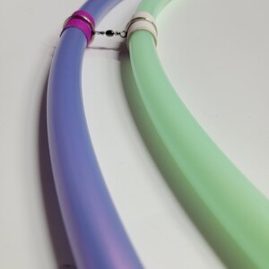 Op de afbeelding: Close-up van twee verbonden, gebogen buizen. Eén buis is een verloop van paars en blauw, de andere is lichtgroen. De buizen zijn verbonden door een kleine metalen draaisluiting en worden bij elkaar gehouden door roze en witte ringen.