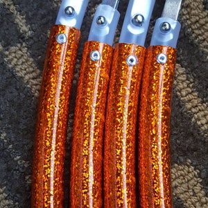 May include: Four orange glitter tubes with silver clasps and clear plastic ends are arranged vertically. Each tube has a small silver rivet. The tubes are on a brown and tan textured surface. The tubes are likely part of a larger product.