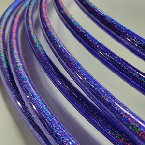 Peut inclure: Gros plan de plusieurs cerceaux violets incurvés avec une finition pailletée et irisée. Les cerceaux sont disposés en parallèle, se chevauchant, créant une sensation de profondeur et de texture. Les paillettes reflètent un arc-en-ciel de couleurs.