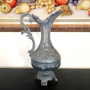 Op de afbeelding: Een kan van tin met een sierlijk ontwerp en een bloemenmotief. De kan heeft een brede basis en een gebogen tuit. Het is een decoratief stuk in vintage stijl.
