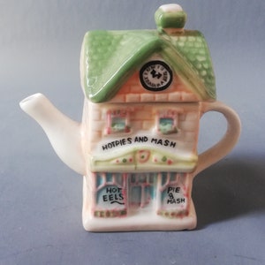 Könnte beinhalten: Eine weiße Keramik-Teekanne in Form eines kleinen Hauses mit grünem Dach. Die Vorderseite der Teekanne trägt ein Schild mit der Aufschrift "Hot Pies and Mash" sowie zwei kleinere Schilder mit der Aufschrift "Hot Eels" und "Pie & Mash".