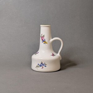 Könnte beinhalten: Ein weißer Keramikkrug mit breitem Boden und engem Hals, der einen einzelnen Henkel auf der rechten Seite aufweist. Die Oberfläche ist mit kleinen, bunten Blumenmustern in Rosa-, Blau-, Gelb- und Grüntönen verziert, die über den Körper verteilt sind.