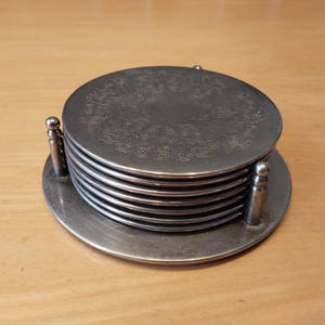 Könnte beinhalten: Ein Set silberfarbener Untersetzer mit einem verzierten floralen Design. Die Untersetzer sind auf einem passenden Sockel gestapelt und werden von zwei kleinen Pfosten gehalten. Die Untersetzer sind aus Metall und kreisförmig.