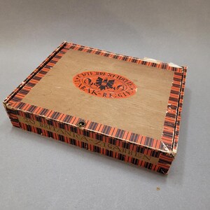 May include: A rectangular wooden cigar box with a light brown top and sides. The top features an oval orange emblem with text and a decorative design. The box sides have a repeating orange and black striped pattern. The text "20 BRITANICA-ZIGARREN" is printed on the side.