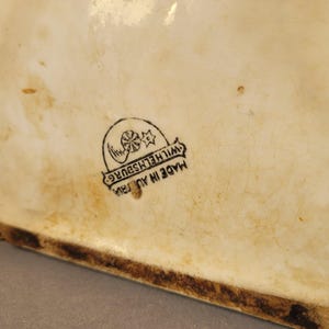 Puede incluir: Pieza de cer&aacute;mica color crema con base marr&oacute;n oscuro. La superficie muestra decoloraci&oacute;n relacionada con la edad y un sello negro que dice "MADE IN AUSTRIA" y "WILHELMSBURG".