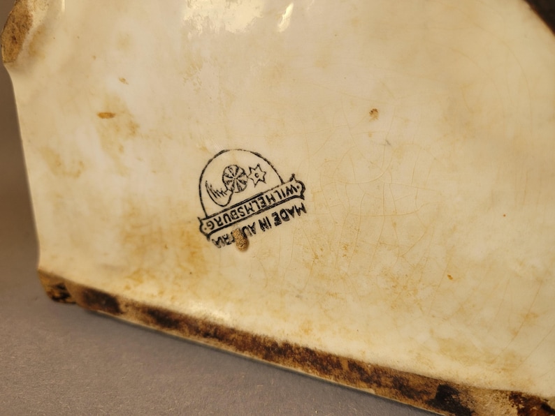 Puede incluir: Pieza de cer&aacute;mica color crema con base marr&oacute;n oscuro. La superficie muestra decoloraci&oacute;n relacionada con la edad y un sello negro que dice "MADE IN AUSTRIA" y "WILHELMSBURG".