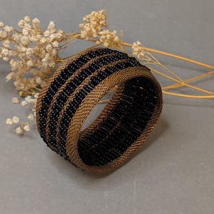 Könnte beinhalten: Ein breites Armband aus metallischem Bronzedrahtgeflecht mit drei markanten horizontalen Streifen aus kleinen, runden, schwarzen Perlen, die in die gewebte Struktur eingearbeitet sind und ein strukturiertes, zylindrisches Design bilden.